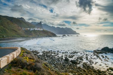 Tenerife 'deki Taganana kıyı manzarası engebeli uçurumları, kayalık kıyı şeridi ve uzak köyüyle. Güneş ışığı okyanusa yansıyarak bulutlu bir gökyüzünün altındaki dramatik manzarayı vurgular.