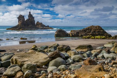Tenerife 'deki Playa Benijo plajında engebeli uçurumlar ve bulutlu mavi gökyüzünün altında hafif okyanus dalgaları. Görkemli kaya oluşumları resim gibi ve sakin bir kıyı manzarası yaratır.