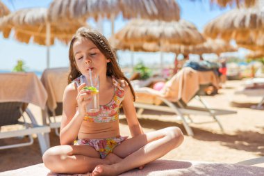 Çiçekli bikinili genç kız güneş şemsiyeleri altında plaj havlusunda çapraz bacaklı otururken samanla alkolsüz mojito içiyor.