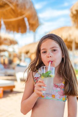 Canlı mayo giymiş rahat genç bir kız plaj şemsiyesinin altında uzanırken pipetten mojito yudumluyor.