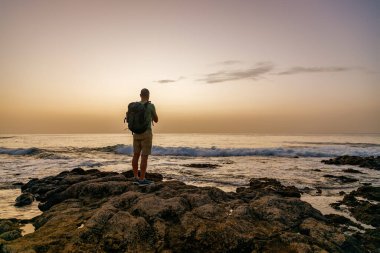 Sırt çantalı yalnız gezgin kayalık sahil şeridinde duruyor Tenerife 'deki macera yolculuğu sırasında Atlantik okyanusu üzerinde altın gün batımını seyrediyor.