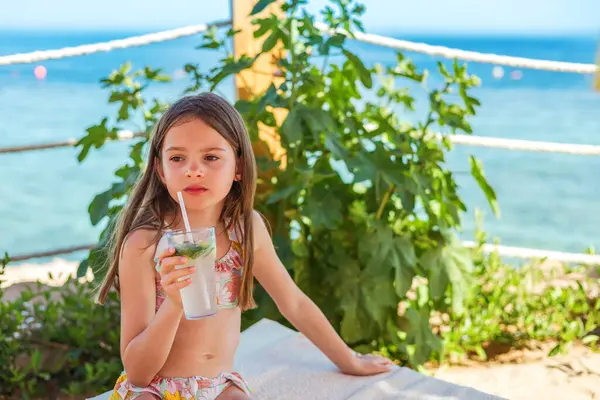 Bir tatil anı yakalandı: Çiçekli bikinili genç bir kız gölgede sakince oturur, güneşli bir ortamda soğuk içkisinin tadını çıkarır.