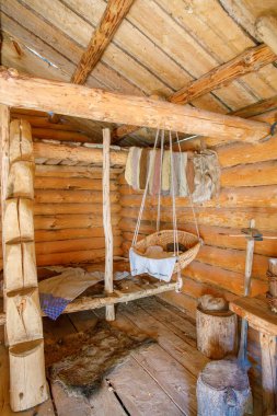 Odunlu ahşap kabin içi, kürk battaniyesi, asılı beşik, iplik demetleri ve rahat el yapımı atmosfer.