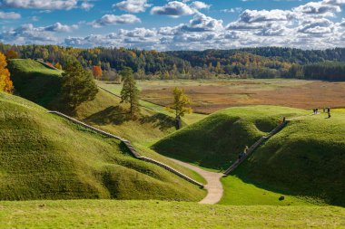 Kernave, Litvanya 'da sakin bir sonbahar manzarası, yumuşak tepeler, kuru otlar ve yumuşak bir uyum içinde güneş ve bulutları harmanlayan bir gökyüzünün altında dolambaçlı bir patika.