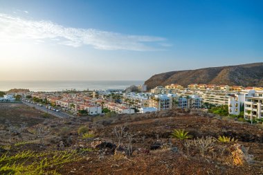 Tenerife 'nin Palm Mar kıyı kasabası, okyanusa doğru uzanan yoğun bir yerleşim alanı dizisini gösteriyor, canlı bir günbatımı gökyüzünün altında engebeli bir tepe ile destekleniyor, seyahat, emlak ve ada hayatı temaları için mükemmel.