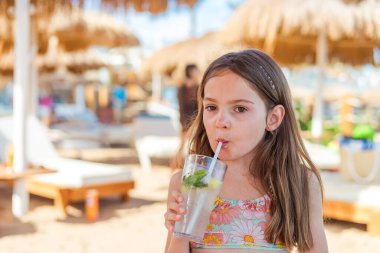 Güneşli bir sahilde ferahlatıcı alkolsüz mojito içen küçük bir kız, başında güneş şemsiyeleri olan çiçekli bir bikini giyiyor.