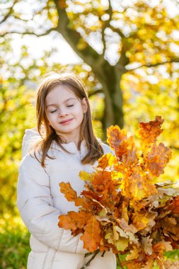 Mutlu genç kız elinde renkli sonbahar yaprakları ile altın güneş ışığı ve arka planda beyaz önlüklü buket tutarak ılık çocukluk anıları yaratıyor.