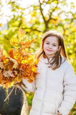 Küçük bir kız, güneşli bir sonbahar gününde elinde kocaman bir buket altın meşe yaprağı ile kameraya gülümsüyor.