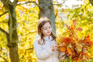 Sonbahar parkında elinde altın meşe yaprakları, canlı mevsimsel renkler ve doğal atmosferle çevrili mutlu genç bir kızın portresi.