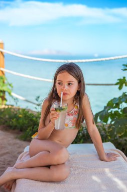 Samimi bir plaj anı, genç bir kızın şemsiye gölgesinin altında ferahlatıcı, alkolsüz bir mojito ile serinlediğini gösteriyor.