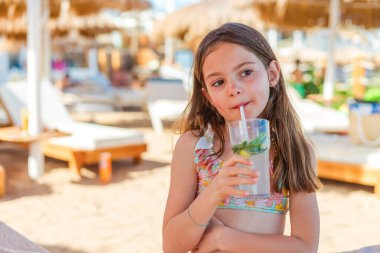 Neşeli bir çocuk plaj şemsiyelerinin altında gevşiyor, tropik bir ortamda soğuk alkolsüz mojito yudumluyor, yazın kaygısız ruhunu somutlaştırıyor.