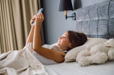 Genç kız rahat bir yatakta uzanıp uyandıktan sonra akıllı telefonunu kontrol ediyor. Modern çocukluk kavramı, teknoloji ve sabah keyfi.