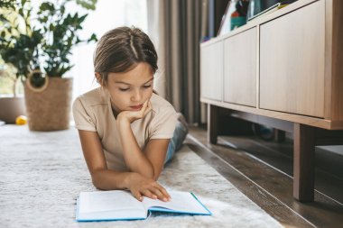 Genç bir kız, parlak, modern bir oturma odasında yerde yatıp kitap okurken, çocukluk eğitimini ve boş zamanlarını temsil eder.
