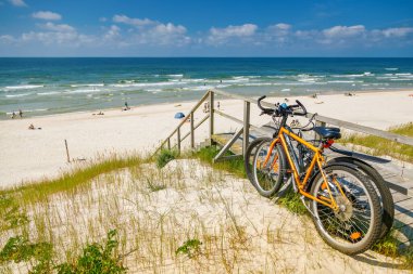 Two orange and black bicycles parked on a wooden ramp leading down to a wide, sunny beach of the Curonian spit, representing summer travel and active vacation