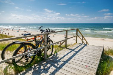 Two bicycles parked on a wooden viewing platform above the wide, sunny Curonian spit beach and Baltic sea, representing active summer travel and leisure