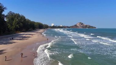 Tayland 'daki tropikal bir plajın havadan 4K görüntüsü. Okyanus dalgaları yavaşça kumlu kıyıya vuruyor. İnsansız hava aracı kıyı şeridinde, mavi suda ve yemyeşil alanda yakalandı.