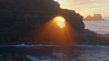 Rays of setting sun break through a hole in the rock off the coast of the Atlantic Ocean on La Palma island, Canary. Great sunset at ocean coast, aerial view. Poris de Candelaria viewpoint, La Palma