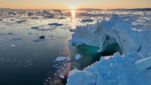 小さなヨットは日没の光の中で氷山の間を帆走する 船はグリーンランドのイルリサットの氷山の巨大な氷河の近くを通過します 北極圏でヨットを旅する 空中撮影 — ストック動画