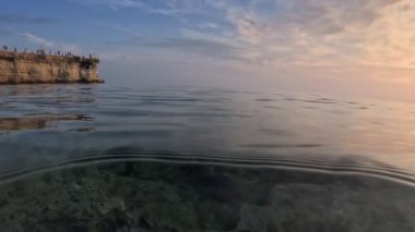 Kıbrıs Rum Kesimi kıyısındaki Akdeniz 'de balık sürüsü. Gün batımında yarı su altında ikiye bölünmüş çekim. Kayalar, berrak deniz ve günbatımı gökyüzü - Kıbrıs 'ın görkemli doğası