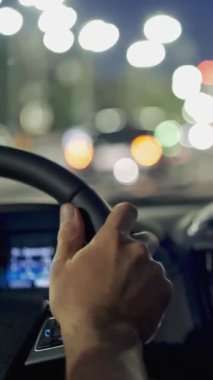 Adam elleri direksiyonda. Akşam kentinde trafik sıkışıklığında araba kullanmak. Gece şehrinden arabayla geçmek. Şehir trafiği ve ışıklar. Dikey çekim