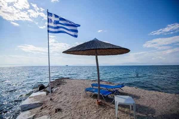 stock image Beach chairs and umbrellas with the national flag of Greece on the sand beach at bay. Beautiful seascape. Blue sky with clouds. Summer travel concept.