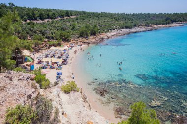 Rahatlatıcı insanlar, turkuaz deniz ve mavi gökyüzü çam ormanlarıyla çevrili bir kumsalda cennet manzarası yaratır. Resimli bir Akdeniz tatil köyünün sakin körfezi.