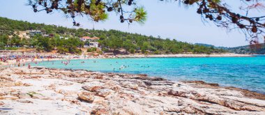 Rahatlatıcı insanlar, turkuaz deniz ve mavi gökyüzü çam ormanlarıyla çevrili bir kumsalda cennet manzarası yaratır. Resimli bir Akdeniz tatil köyünün sakin körfezi.