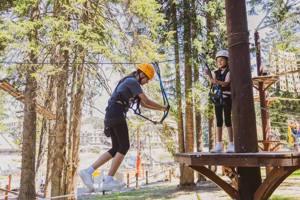 Girl crosses a challenging rope course in a forest at amusement park, wearing protective gear.