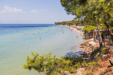 Turistlerin berrak mavi sularda dinlenip yüzdüğü canlı plaj sahnesi.