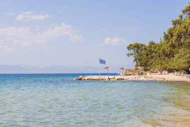 Sahil boyunca Yunan bayrağı ve güneş şemsiyeleriyle huzurlu sahil sahnesi.