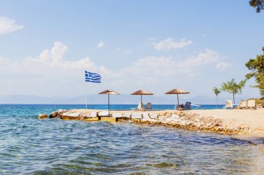 Sahil boyunca Yunan bayrağı ve güneş şemsiyeleriyle huzurlu sahil sahnesi.