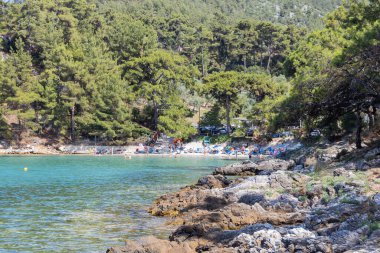 Ağaçlarla çevrili, arka planda turistler olan berrak mavi suyu olan gizli kayalık plaj..