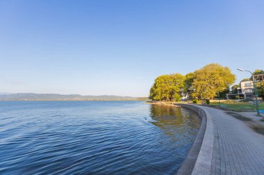 Doiran Gölü 'nün kıyısında, sakin suların günbatımının ılık ışığını yansıttığı engebeli bir göl yolu. Kuzey Makedonya