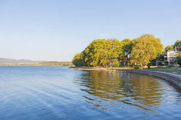 Doiran Gölü 'nün kıyısında, sakin suların günbatımının ılık ışığını yansıttığı engebeli bir göl yolu. Kuzey Makedonya