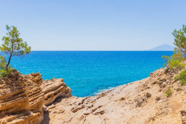 Kayalık kayalıklar, güneşli bir yaz gününde, çam ağaçlarının çerçevelediği canlı turkuaz denizle karşılaşıyor..