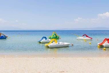 Sakin, kristal berrak sularda yüzen renkli pedallı teknelerin olduğu sakin sahil sahnesi.