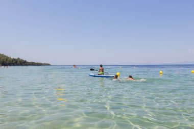 Çocuklar sığ sularda sığ bir yaz gününün tadını çıkarırken, diğerleri kıyı şeridi yakınlarındaki ferahlatıcı denizde yüzerken biri kürek tahtasının üzerinde..