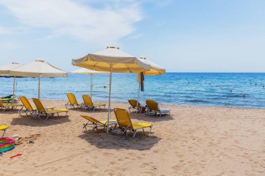 Kumlu bir sahilde, parlak sarı güneş şezlongları ve beyaz şemsiyeler, rahatlatıcı bir sahil kaçışı sunuyor..