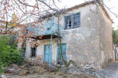 Eski, terk edilmiş bir ev solmuş mavi panjurlar ve çökmüş bir cephe, geleneksel bir Yunan köyünde vahşi yeşilliklerle çevrili..