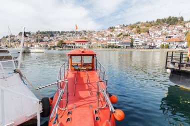 Canlı kırmızı tekne Ohri Gölü kıyısına yanaşırken, arka planda resmedilmiş Ohri kasabası yer alıyor..