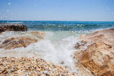 Sakin kayalık bir plajın, pürüzsüz çakıl taşları ve berrak mavi suyla resimli bir sahnesi..
