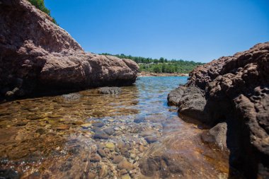 Sakin kayalık kıyı şeridinde kristal berrak suyun altında pürüzsüz çakıl taşları var. Çerçevelenmiş engebeli kayalar ve parlak mavi bir gökyüzü.