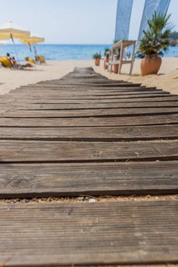 Kırsal ahşap kaldırım, arka planda güneş şemsiyeleri ve deniz olan kumlu bir sahile doğru uzanıyor..