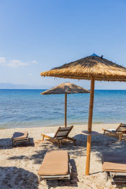 Saman şemsiyeler ve ahşap güneşlikler sakin mavi denize bakan kumlu bir plaja saçılmış..