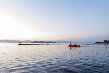 Gün batımında sakin bir denizin sakin manzarası, küçük kırmızı bir tekne suda süzülüyor. Gökyüzü yumuşak pembe ve turuncu renklerle boyanır. Huzurlu ve resimsel bir atmosfer yaratır..