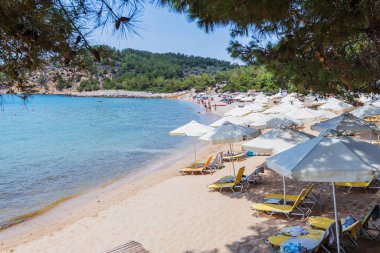 Güneşli şezlonglar, şemsiyeler ve kristal berrak turkuaz sularla sakin sahil sahnesi, Yunanistan 'da rahatlatıcı bir yaz tatili için mükemmel..