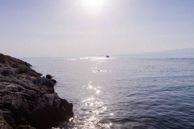 Ufukta süzülen küçük bir teknesi olan, güneşin sıcacık parıltısıyla yıkanan sakin bir deniz manzarası. Kristal berraklığında su, güneş ışığını yansıtarak büyüleyici bir desen oluşturur..