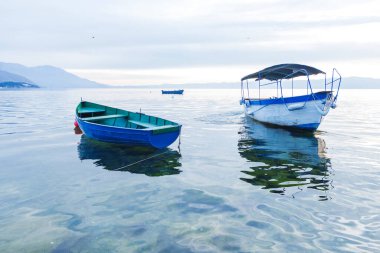 Sakin göl sahnesinde sakin, berrak sularda yüzen tekneler ve arka planda bulutlu bir gökyüzü var..
