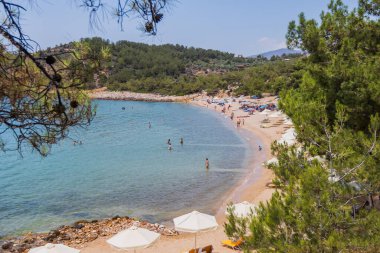 Güneşli şezlonglar, şemsiyeler ve kristal berrak turkuaz sularla sakin sahil sahnesi, Yunanistan 'da rahatlatıcı bir yaz tatili için mükemmel..
