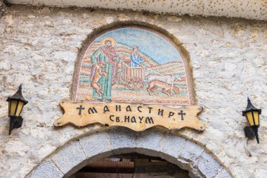 Ohri Gölü 'ndeki St. Naum Manastırı girişinin üzerinde yer alan dini bir sahneyi resmeden renkli bir mozaiğin yakın görüntüsü. Mozaiğin karmaşık detayları ve canlı renkleri manastırın zengin tarihini ve sanatsal mirasını vurguluyor..